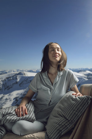 A young woman in pajamas with her eyes closed in bed against the background of snowy mountains. A cozy, peaceful atmosphere of nature.の写真素材