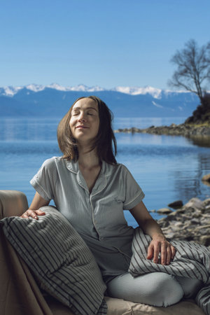 A young beautiful woman in pajamas with her eyes closed in bed against the background of a lake and snowy mountains. A cozy, peaceful atmosphere of nature.の写真素材