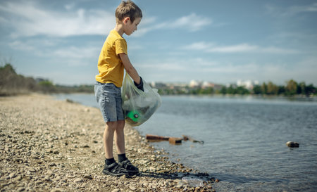 A child boy volunteer in gloves tidying up rubbish plastic garbage from the riverbank. Children take care of nature.の写真素材