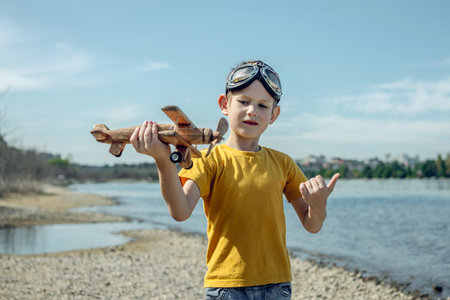 A child boy runs with a wooden retro airplane wearing pilot glasses. Dreams of freedom and travel. Playing against a blue sky background.の写真素材