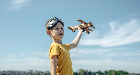 A child boy runs with a wooden retro airplane wearing pilot glasses. Dreams of freedom and travel. Playing against a blue sky background.の写真素材