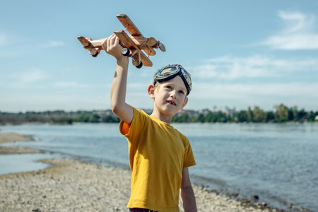 A child boy runs with a wooden retro airplane wearing pilot glasses. Dreams of freedom and travel. Playing against a blue sky background.の写真素材