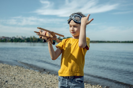 A child boy runs with a wooden retro airplane wearing pilot glasses. Dreams of freedom and travel. Playing against a blue sky background.の写真素材