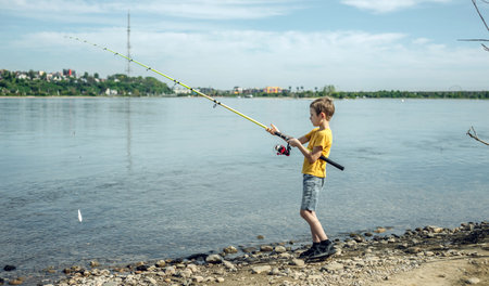 A child boy with a fishing rod is fishing standing on the riverbank. Active children's recreation on a sunny summer day.の写真素材