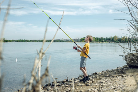 A child boy with a fishing rod is fishing standing on the riverbank. Active children's recreation on a sunny summer day.の写真素材