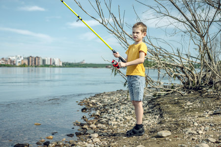 A child boy with a fishing rod is fishing standing on the riverbank. Active children's recreation on a sunny summer day.の写真素材