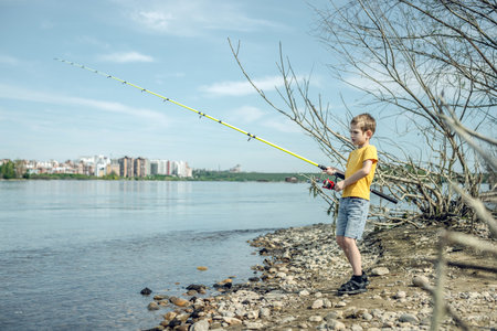 A child boy with a fishing rod is fishing standing on the riverbank. Active children's recreation on a sunny summer day.の写真素材