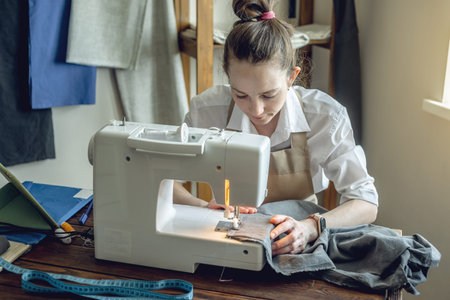 A female tailor is working on a sewing machine stitching fabric in sunny room. Home sewing...