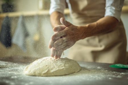 A baker in an apron kneads the dough with his hands and sprinkles it with flour to bake delicious...