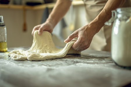 A baker in an apron kneads the dough with his hands and sprinkles it with flour to bake delicious homemade bread. Rustic style on a dark backgroundの写真素材