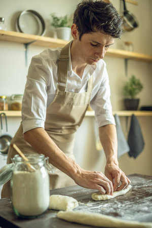 A baker in an apron kneads the dough with his hands and sprinkles it with flour to bake homemade...