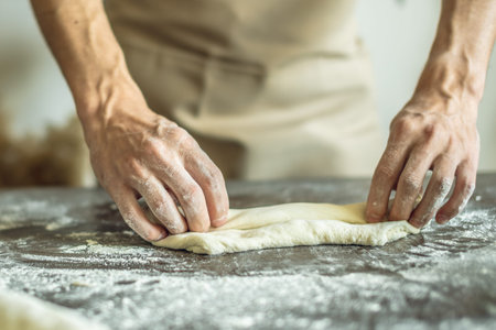 A baker in an apron kneads the dough with his hands and sprinkles it with flour to bake delicious...
