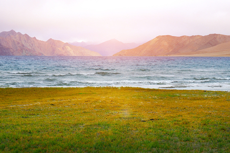 Colors of Pangong Lake, scenic view of Pangong Tso against the background of distant mountain range and cloudy blue sky with green field, Ladakhの写真素材