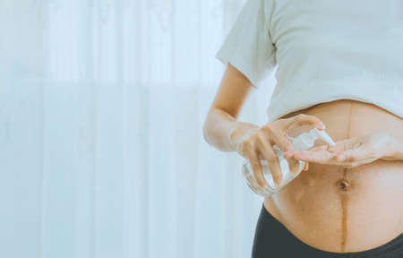 A pregnant woman wears a surgical mask is washing hands by alcohol gel on the sofa. Protect a COVID-19 (Coronavirus), PM 2.5 and prevent infection to the fetus concept.の写真素材