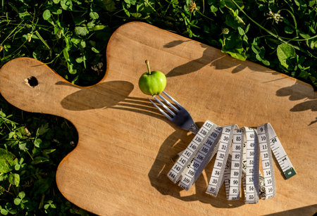 An apple on a fork on a cutting board, measuring tape, against a background of green grass. Diet...