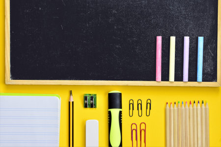 Varied school supplies from small blackboard with chalks and daily supplies at school, on yellow...