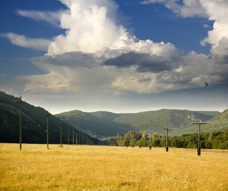 Power line is located in a mountain valley in the pasture with dry grass on a background of white large clouds.の写真素材