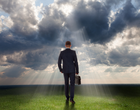 Confident businessman with briefcase moves towards of dramatic clouds on the green grass, business concept.の写真素材