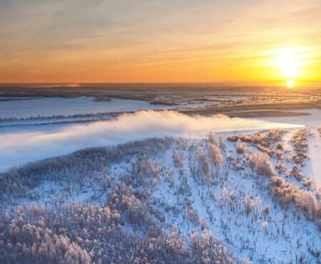 The Aerial view the river on snow-covered forest plain in time of cloudy winter evening. Crowns of coniferous trees are lighted up by a bright setting sun.の写真素材