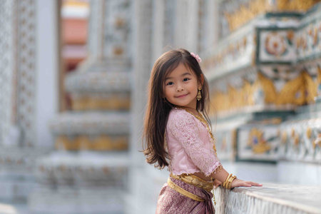 A cute young girl in a shimmering traditional Thai outfit smiles sweetly at the camera.の素材