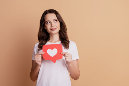 Portrait of dreamy curious girl hold notification heart icon look empty space isolated on beige...