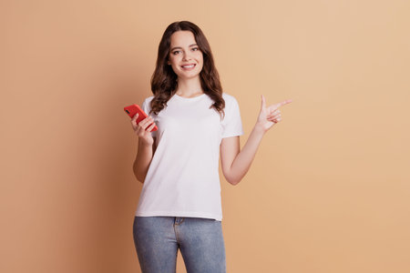 Photo of pretty promoter lady hold phone indicate forefinger empty space posing on beige background