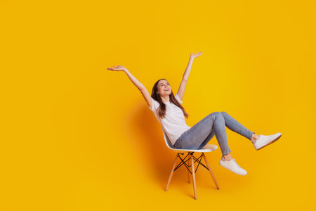 Photo of dreamy inspired lady sit chair raise arms look up empty space wear white t-shirt posing on yellow backgroundの写真素材