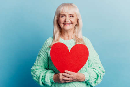 Smiling elderly woman hold red heart card isolated on blue backgroundの写真素材
