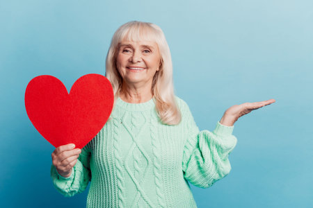 Smiling elderly woman hold hand advertise red heart card isolated on blue background