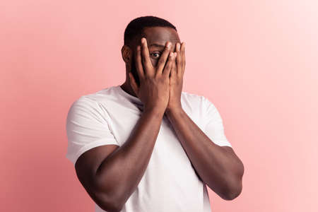 Headshot of young shy man cover face palms peeking through fingers on pink wallの写真素材