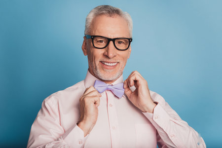 Photo of attractive aged man adjusting bow tie isolated on blue background.