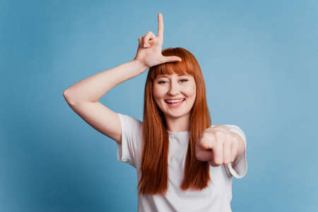 Photo of rude young girl showing lose sign point finger at camera isolated over blue backgroundの写真素材