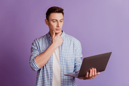 Portrait of minded focused concentrated guy hold laptop finger chin on violet background