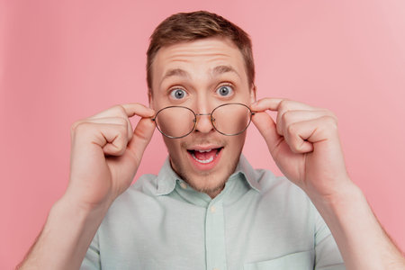 Photo of young handsome man amazed shocked surprised hands touch glasses isolated over pink color...