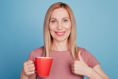 Photo of cheerful woman holding mug of coffee show thumb-up like cool advert isolated over blue...