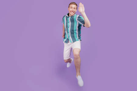 Portrait of friendly positive cheerful guy jump run wave hand on violet backgroundの写真素材