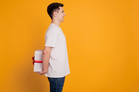 Profile portrait of funny guy hold gift package behind back look empty space make surprise on yellow...
