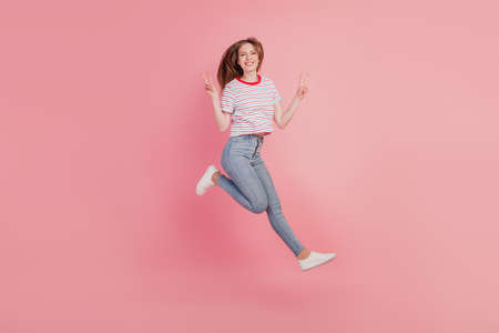 Portrait of cheerful carefree lady jump run have fun show v-signs on pink backgroundの写真素材
