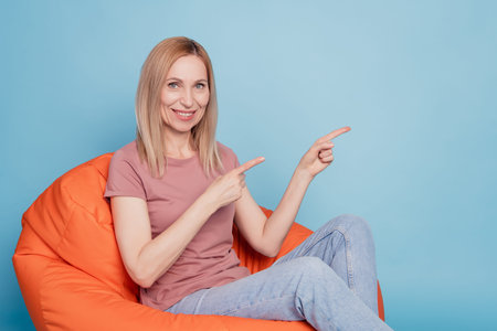 Full size photo of happy woman indicate fingers empty space sit chair bean wear t-shirt advert...