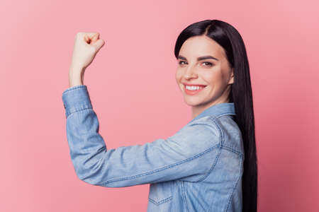 Profile side view portrait of cute cheerful girl showing strong arm muscle isolated on pink pastel color backgroundの写真素材