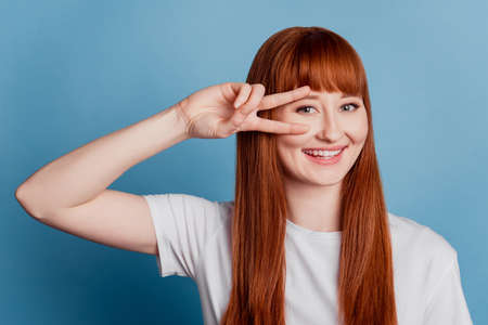 Portrait of cheerful girl showing v-sign near eyes isolated on blue backgroundの写真素材