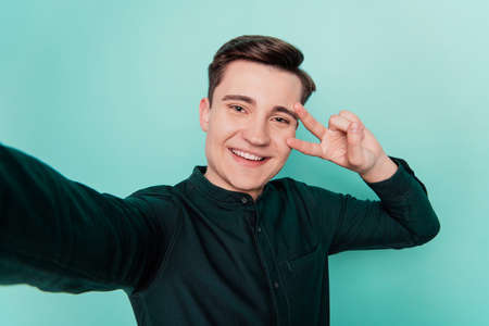 Portrait of friendly man shoot selfie shiny white smile show v-sign cover eye on turquoise backgroundの写真素材