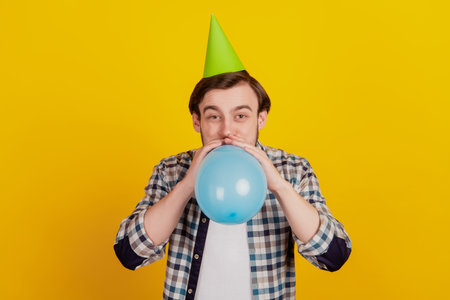 Photo of young guy blow balloon wear paper cone hat birthday party decoration isolated yellow color backgroundの写真素材