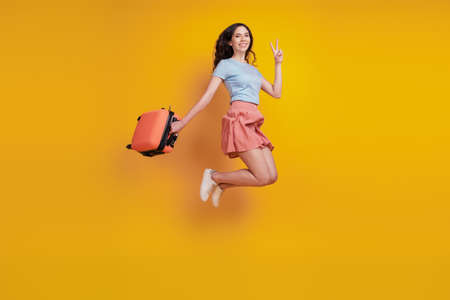 Full length photo of young woman happy positive smile bag suitcase jump show peace v-sign isolated over yellow color backgroundの写真素材