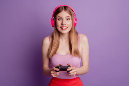 Portrait of cheerful carefree lady hold controller play video game wear earphones pink top on violet backgroundの写真素材