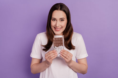 Portrait of a happy young cheerful woman eat food calories chocolate bar isolated over violet backgroundの写真素材
