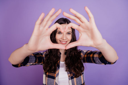 Cute beautiful girl in love making heart with fingers and smiling romantic isolated over violet...