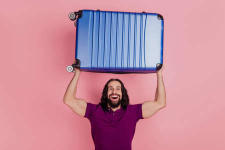 Photo of excited amazed man hold valise raise hands up wear purple t-shirt isolated on pink color backgroundの写真素材