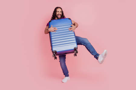 Photo of carefree funky man cuddle valise dance wear purple t-shirt isolated on pink color backgroundの写真素材