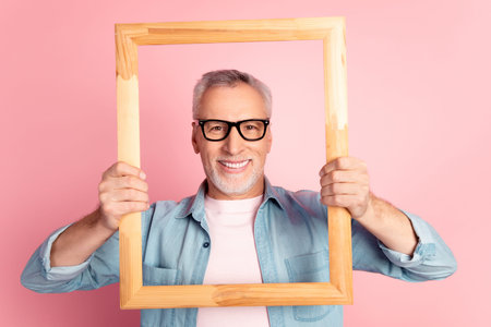 Photo of cheerful aged man hold wooden frame make picture eyewear isolated over pink color...